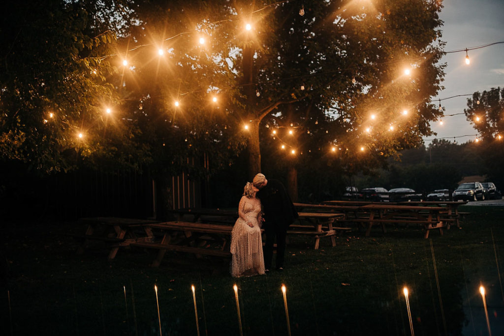 bride and groom kiss on picnic tables under twinkle lights