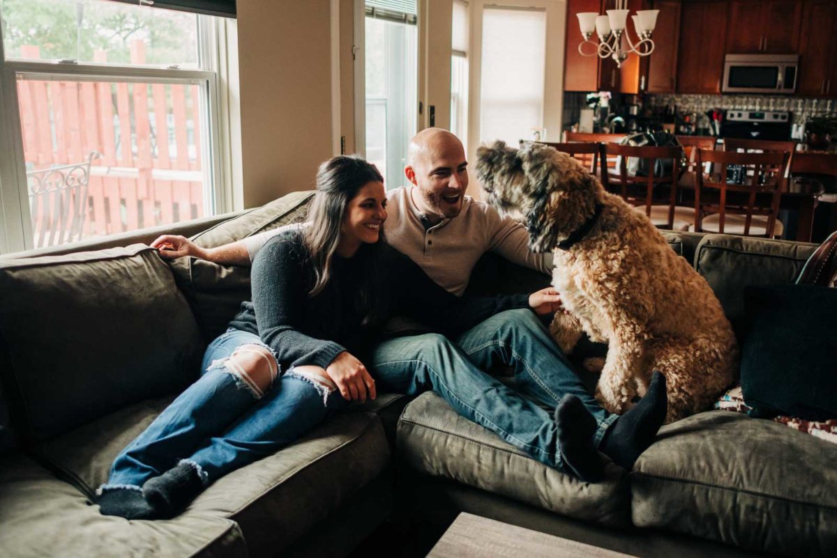 An engaged couple plays with their dog on their couch